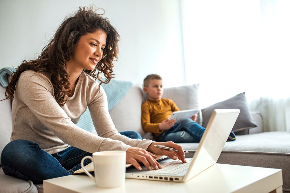 Image of a woman on her computer with her child on the couch in the background